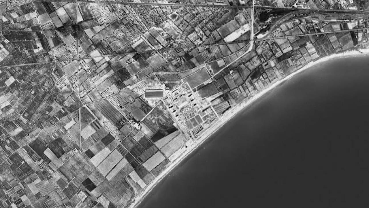 Czarno-biała, pozioma fotografia z perspektywy lotu ptaka. Krajobraz przybrzeżny z widocznymi drogami, polami uprawnymi oraz budynkami.